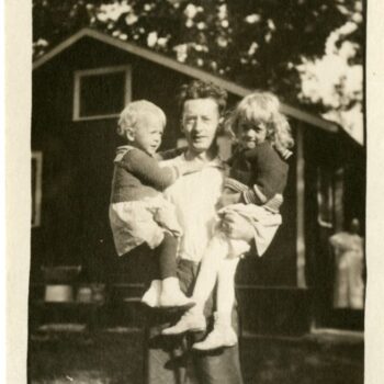 Young man holds two small children with house in background.
