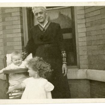 Grandma Johnson poses with two children. One in highchair and one standing in front.