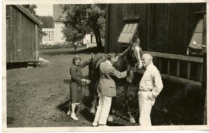 Man and woman groom horse on homestead. One man stand to the right.