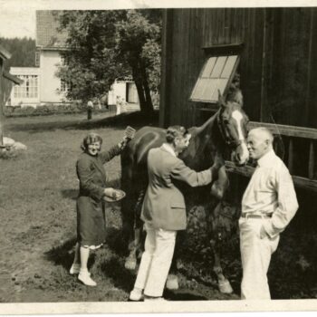 Man and woman groom horse on homestead. One man stand to the right.