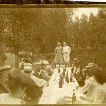 About sixteen people sit around a table outside. Two young children are standing on the end of the table, posing for the photo.