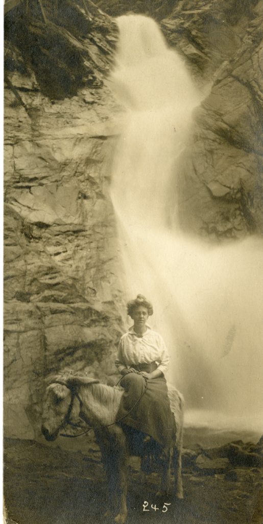 A woman, Nora Bernts, sits on a donkey in front of a waterfall.