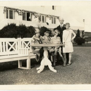 Five individuals outside a house. Three sit on a bench while two stand beside.