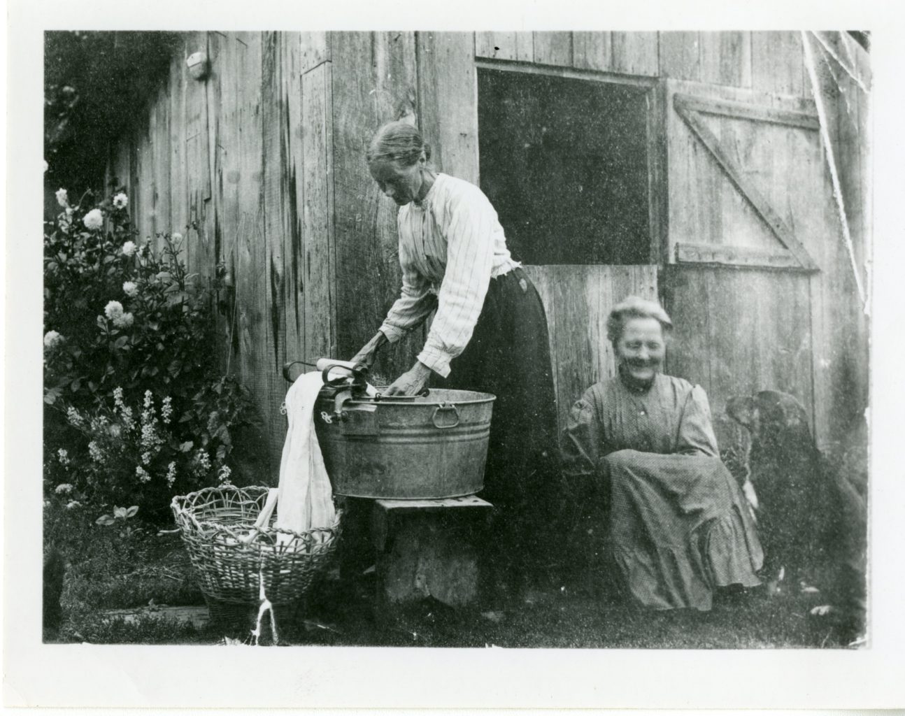 Mrs. Bruflodt and Anne Gausta outside of farm. Mrs. Bruflodt washes clothes, while Anne sits beside with dog.