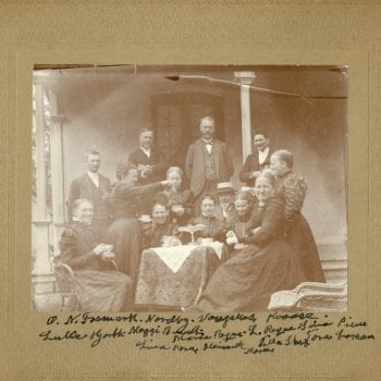 Approximately thirteen adults sit outside around a table drinking coffee.
