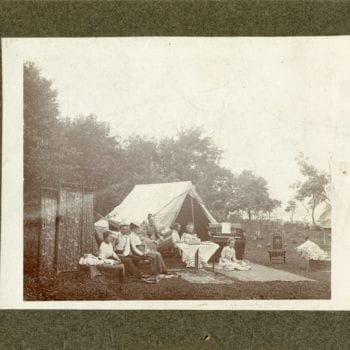 Five women and two men sit outside with tent and rugs. Some holding rackets.