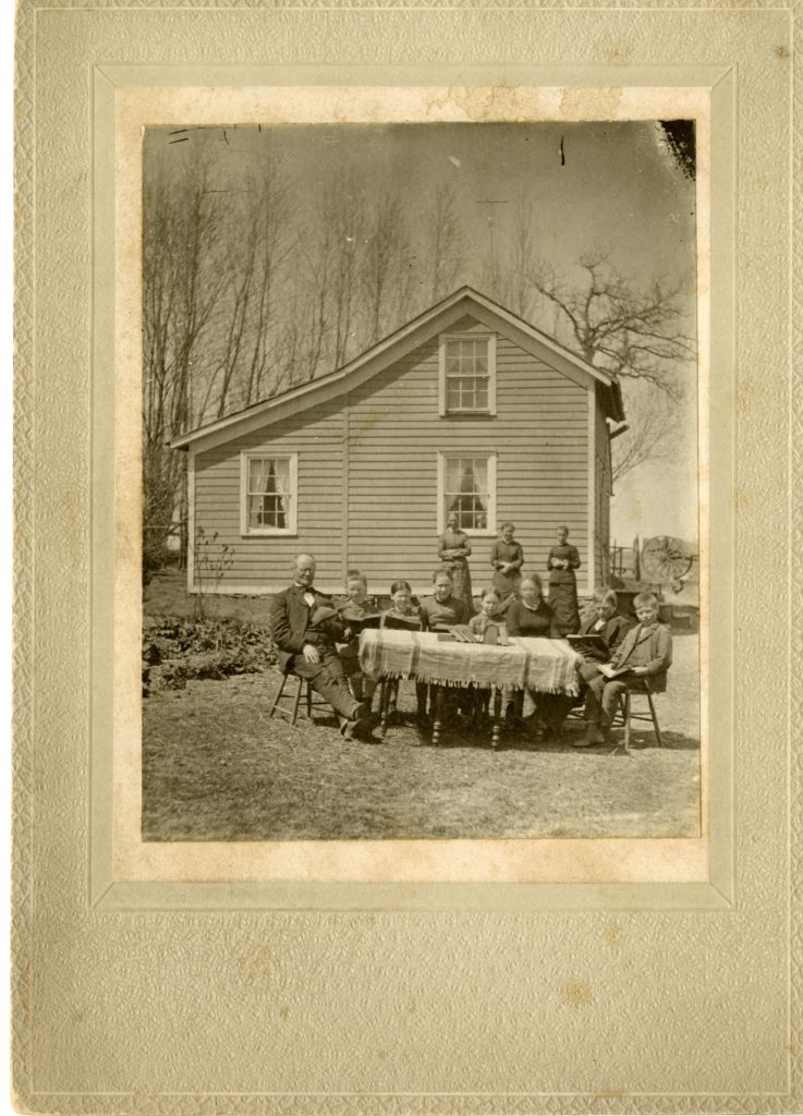 A man and seven children sit around a table outside and read. Three women stand behind and house in background.