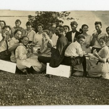 Group of young adults eat around a table outside by the water.
