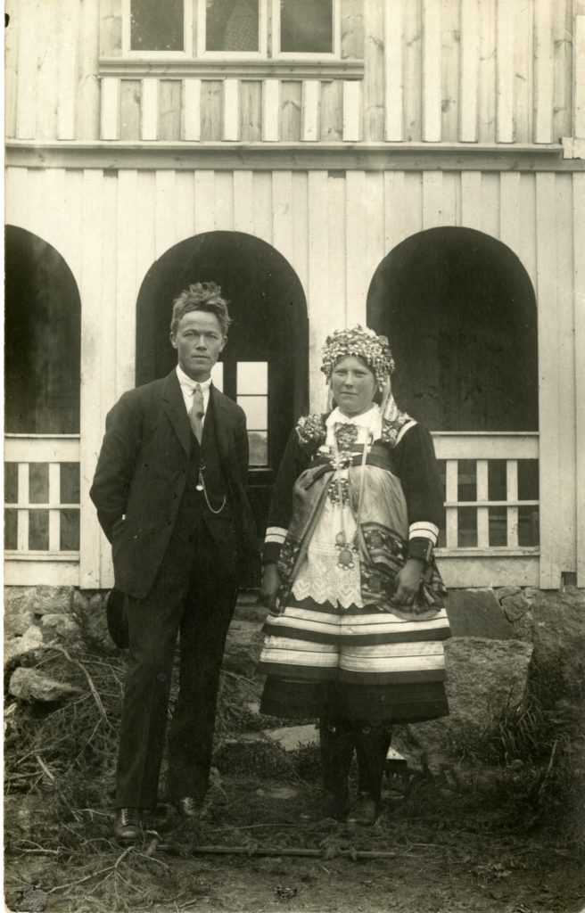 A man and woman pose in front of building. Woman is in traditional Setesdal bunad.
