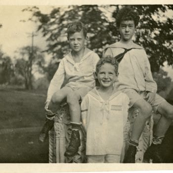 Three boys pose for a photo, two sitting and one standing. All in white.