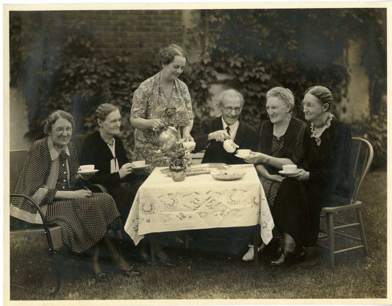 Five women and on man gather for tea outside around a table.