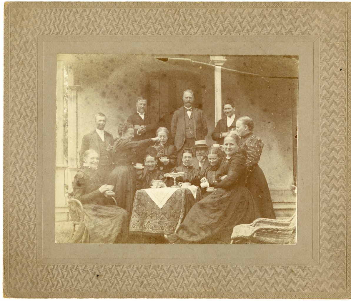 Small group of men and women gather for tea outside around table.