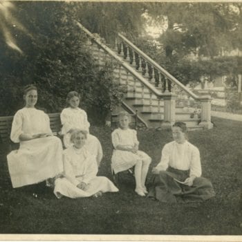 Five young girls sit in a lawn outside of a house.