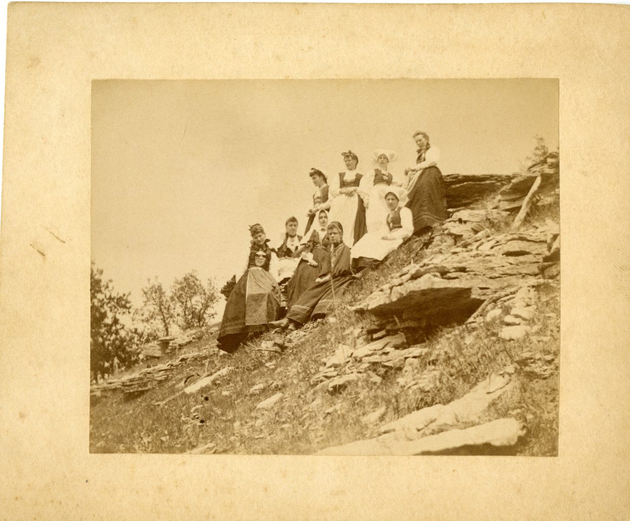 Group of nine women sitting/standing on a hill in Norwegian national dress.