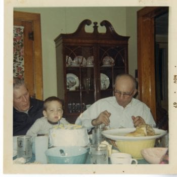 Two older men and young boy sit at the dinner table, the boy blows out a candle on a cake.