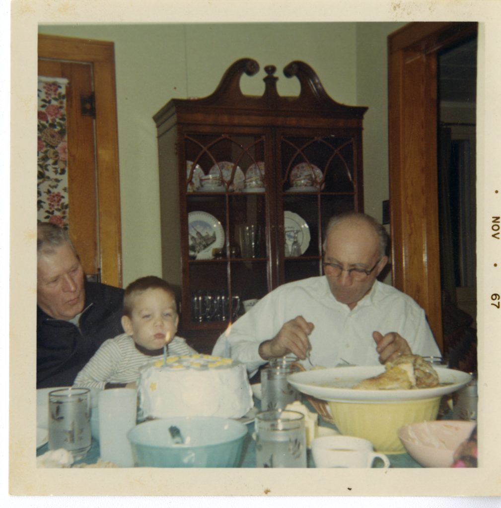 Two older men and young boy sit at the dinner table, the boy blows out a candle on a cake.