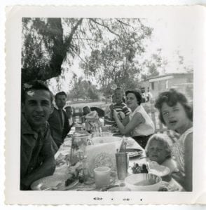 Family of nine sit around a table outside for picnic.