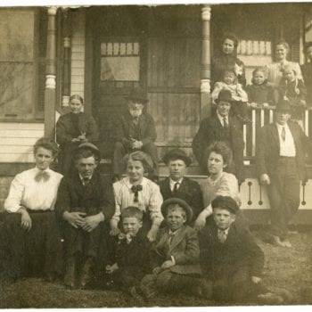 Group of men, women, and children sit outside of a house together.